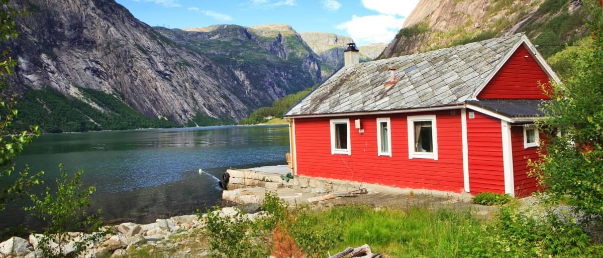 Ein idyllisches rotes Holzhaus an einem See inmitten von Bergen und strahlend blauem Himmel.