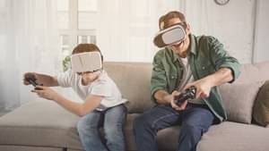 Vater und Sohn sitzen auf der Couch und spielen ein Spiel auf dem Fernseher. Beide haben einen Joystick in der Hand und tragen eine  AR-Brille.