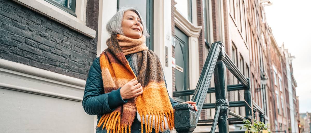 Ältere Frau mit Steppjacke steht auf der Treppe vor ihrem Hauseingang und wirft sich einen gemütlichen Schal um.
