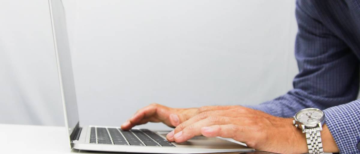Die Hände eines Mannes liegen auf der Tastatur eines Laptops. Daneben liegt ein Smartphone und ein Stift.