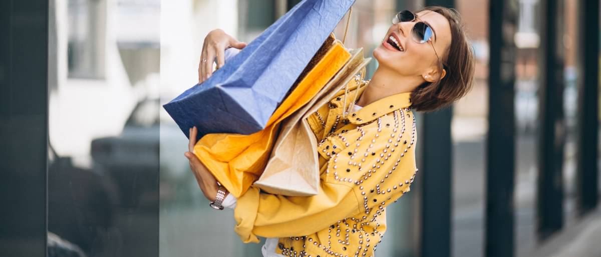 Eine lächelnde junge Frau mit Sonnenbrille geht mit einigen Shoppingtüten an einer Reihe von Schaufenstern vorbei