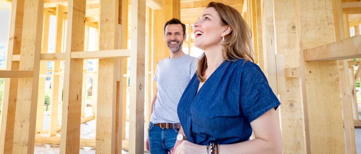 Ein Paar steht in dem Holzgerüst ihres Neubaus. Der Mann schaut lächelnd seine Frau an, die staunend herumschaut.