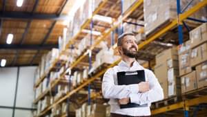 Mann mit Klemmbrett steht vor einem großen Regal in einer Lagerhalle.