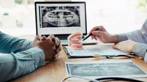 Jemand zeigt mit einem Kuli auf ein Gebiss, im Hintergrund sieht man ein Röntgenbild