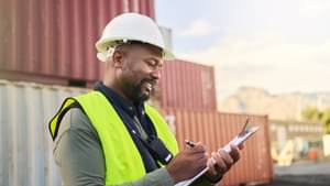 Ein Mann mit gelber Warnweste steht vor Containern und schreibt lächelnd auf einem Klemmbrett