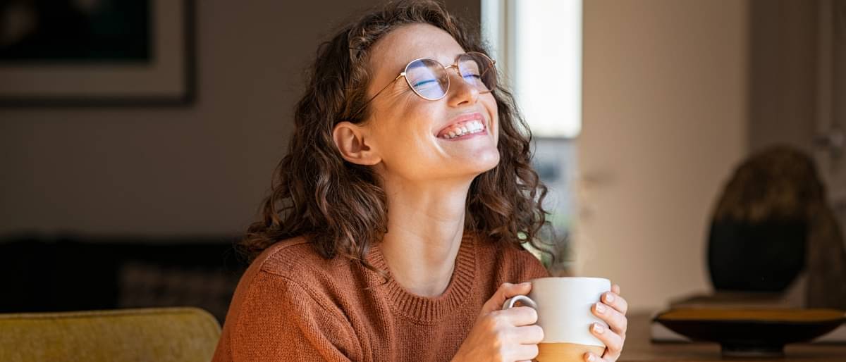 Eine junge Frau lächelt fröhlich, hat die Augen geschlossen und genießt den Augenblick und hat eine Tasse Kaffee oder Tee in beiden Händen.