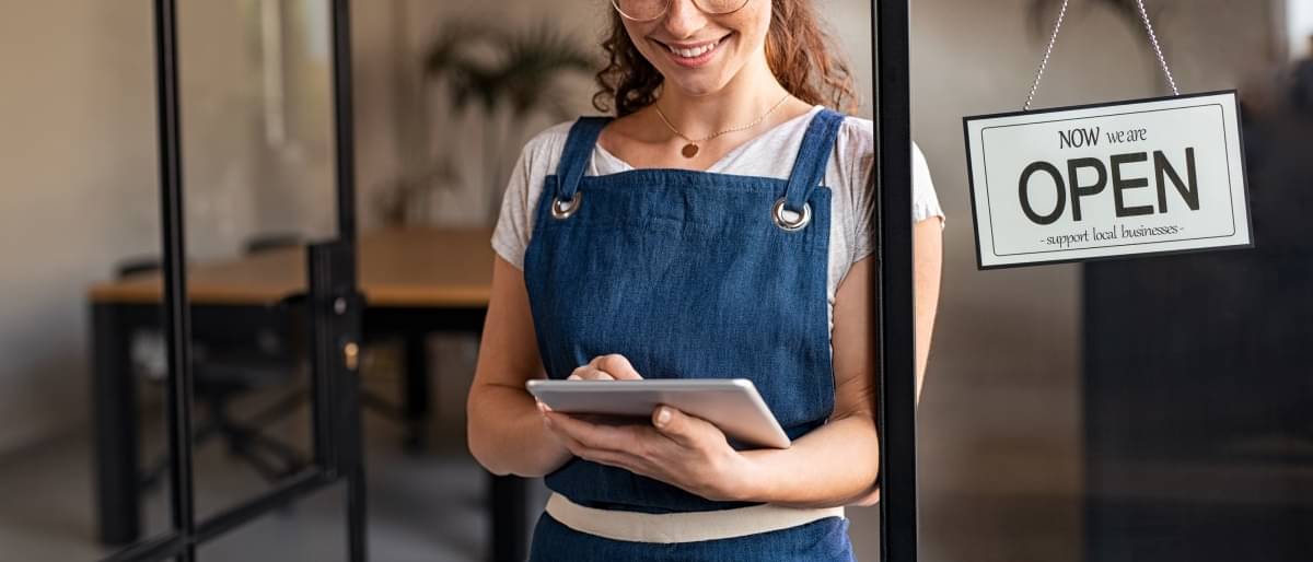 Junge Frau mit Schürze steht in der Tür eines Cafés und schaut glücklich auf ein Tablet.