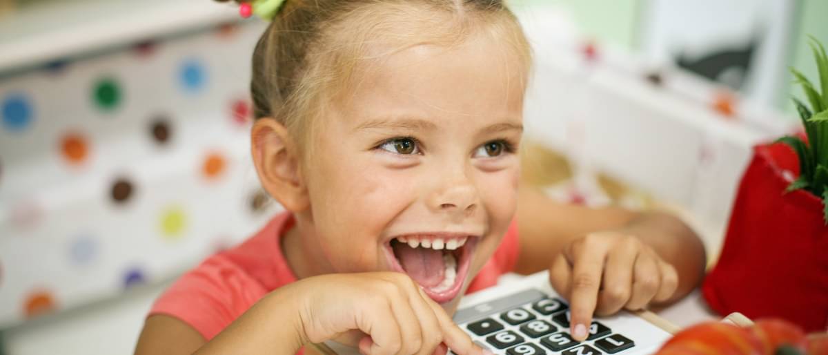 Ein kleines blondes Mädchen mit zwei lustigen Zöpfen spielt herzlich lachend an einer Kinderkaufmann-Kasse.  Im Hintergrund sieht man eine bunt gepunktete Tapete.