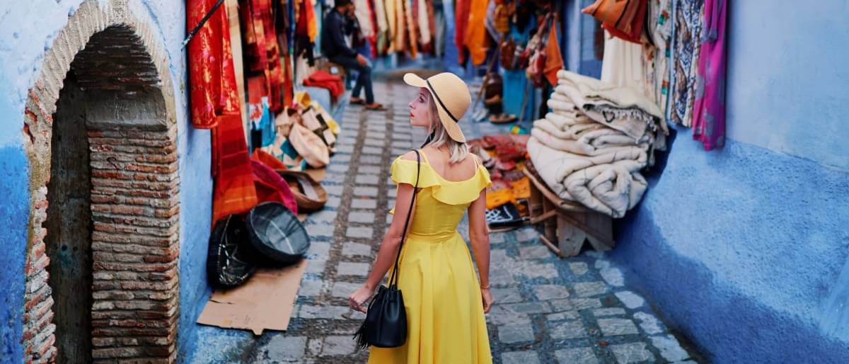 Auf dem Bild ist eine marokkanische Straße zwischen zwei blauen Häuserwänden zu sehen. An den Wänden hängen bunte Tücher. Eine Frau in gelben Kleid und Hut geht die Straße hinauf - Reisetipps Marokko
