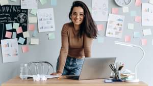 Junge Frau stützt sich lässig auf den Schreibtisch in ihrem Büro und lächelt in die Kamera. Im Hintergrund hängen viele bunte Post-Its an der Wansd.