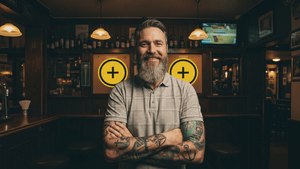 Ein bärtiger Mann mit tätowierten Armen steht lächelnd mit verschränkten Armen in einem warm beleuchteten Pub vor einer holzvertäfelten Wand mit zwei runden gelben Tafeln im Hintergrund.
