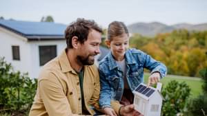 Junger Mann steht mit seiner Tochter im Garten mit einem Modellhaus in der Hand und zeigt auf eine Photovoltaik-Anlage auf dem Dach.