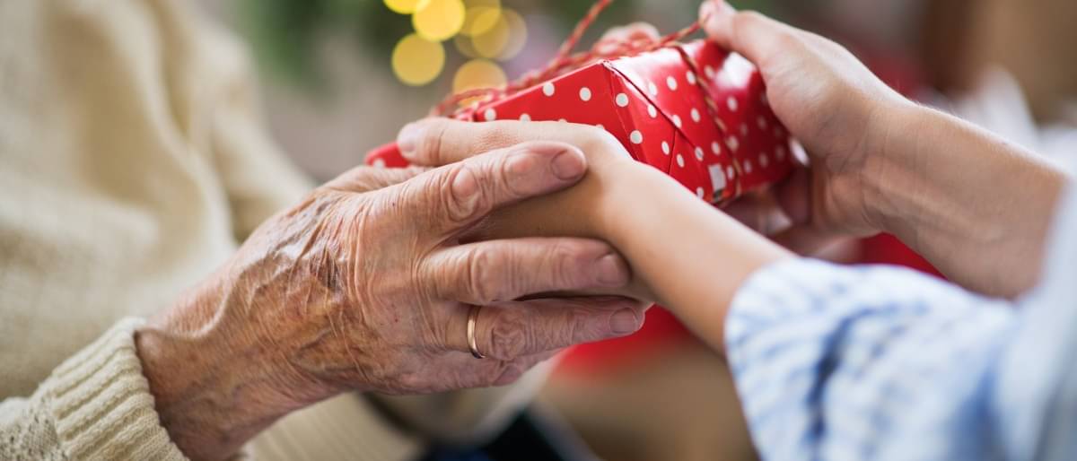 Eine Großmutter reicht ihrem Enkel ein kleines Geschenk, das mit rotem weiß gepunkteten Geschenkpapier eingepackt wurde.