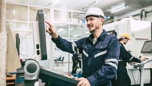 Junger Mann in Overall und Helm steht einer Werkstatt am Bedienelement einer Maschine.