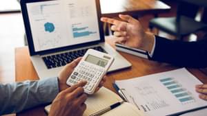 Man sieht die Arme zweier Männer in Business-Kleidung auf einen Tisch gestützt. Um sie liegen einige Unterlagen und ein Laptop steht daneben. Einer der Männer hält einen Taschenrechner in der Hand.