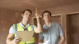 Ein Bauleiter mit einer Warnweste und ein weiterer Mann stehen in einem entkernten Haus und besprechen sich. Der Bauleiter zeigt dabei an die die Decke.