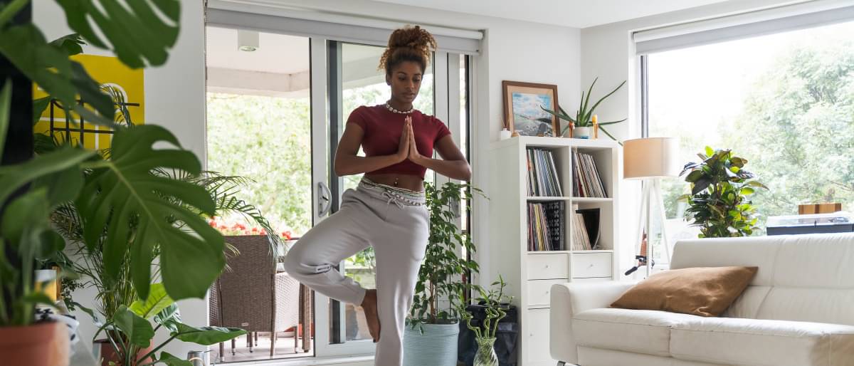 Eine junge Frau steht in einer Yoga-Position auf einem Bein und vor der Brust zusammengeführten Handflächen sowie geschlossenen Augen in Ihrem hellen, weiß möbiliertem Wohnzimmer.