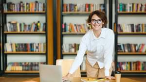 Eine Frau steht glücklich hinter ihrem Schreibtisch, auf dem ein Laptop, mehrere Papiere, Notizbücher und ein Handy liegen. Im Hintergrund erkennt man Regale mit vielen Büchern.