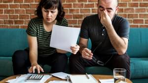 Eine Frau und ein Mann sitzen neben einander, die Frau benutzt einen Taschenrechner, der Mann wirkt sehr gestresst und unglücklich