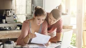Ein junges Paar schaut gemeinsam auf einige Rechnungen, die auf einem Esstisch ausgebreitet sind. Vor ihnen steht ein aufgeklappter Laptop.