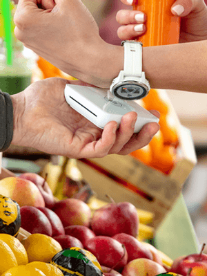 Eine Frau trägt eine weiße Smartwatch. In einem Obst- und Gemüseladen hält sie diese an ein mobiles Bezahlterminal um ihren Einkauf mit Garmin Payzu bezahlen.