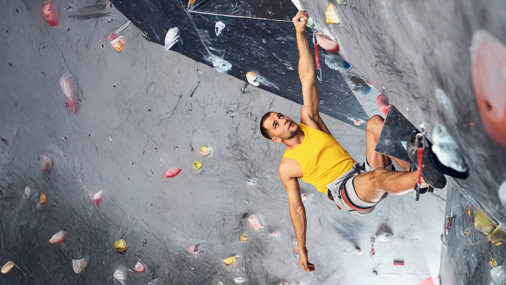 Ein Kletterer mit gelben ärmellosen Shirt hängt mit einer Hand an einer überhängenden Boulderwand..