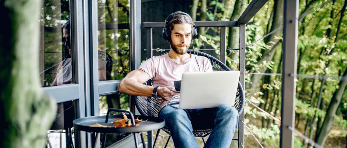 Junger Mann sitzt mit Laptop im Schoß auf Terrasse im Grünen und tippt auf Tastatur.