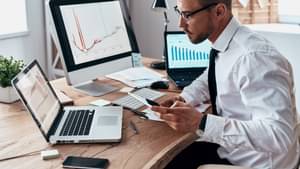 Junger Mann in Hemd und Krawatte sitzt mit seinem Handy in der Hand am Schreibtisch und betrachtet Diagramme auf seinem Laptop.