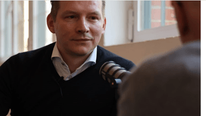 Kay Theuer schau kritisch nach vorne und spricht in ein Mikrofon rein, das vor ihm von einer zweiten Person gehalten wird, die jedoch nur am Rand verschwommen zu sehen ist. 