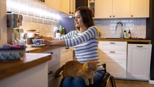 Eine junge Rollstuhlfahrerin mit langen, braunen Haaren macht sich an ihrer Arbeitsfläche in der Küche einen Tee. Auf ihren Oberschenkeln liegt eine Katze mit rot-getigertem Fell.