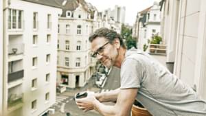 Mann steht am Balkon und lehnt mit Kaffee-Becher in der Hand auf dem Geländer in Altbaustraße.