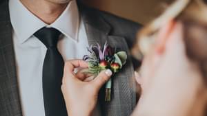 Eine Braut befestigt ein kleines Blumen-Geflecht am Anzug ihres Bräutigams.