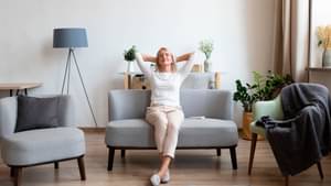 Eine Frau sitzt auf einem Sofa und legt die Hände in den Nacken und schaut sehr zufrieden und entspannt aus.