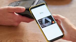 In einem Café wird ein Smartphone zum Bezahlen an ein Kartenlesegerät gehalten. Auf dem Display sind das Google Pay Logo sowie eine schwarze Premium Kreditkarte der Commerzbank zu sehen.