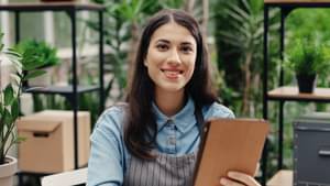 Eine junge Frau lächelt in die Kamera. Sie hat eine Schürze um und steht in einem Café. 