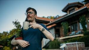 Mann mit Kaffeetasse und Bäumchen im Arm steht vor seinem Haus.