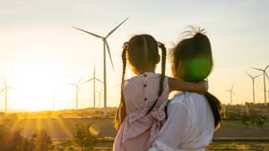 Eine junge Frau hält ein junges Mädchen auf dem Arm. Sie stehen mit dem Rücken zum Betrachter und schauen auf einige Windräder während die Sonne untergeht.