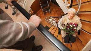 Eine ältere Frau sitzt auf einem Treppenlift und hat einen Strauß Blumen in der Hand