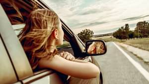 Junge Frau mit blonden Haaren sitzt auf der Beifahrerseite eines Autos und lehnt sich mit Arm und Kopf zum offenen Fenster raus.