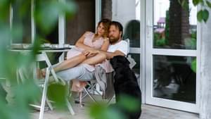 Mann sitzt auf Terrasse im Grünen. Frau sitzt auf seinem Schoß. Hund sitzt daneben auf dem Boden.