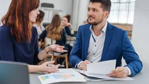 Eine junge Frau befindet sich im Beratungsgespräch mit einem jungen Mann im Anzug. Vor ihnen liegen einige Dokumente.