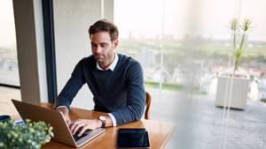 Mann sitzt im Büro am Schreibtisch mit einem vor sich stehendem aufgeklappten Laptop und schreibt konzentriert auf der Tastatur.