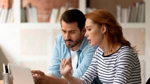 Eine junge Frau und ein junger Mann sitzen an einem Laptop und unterhalten sich über mögliche Kreditsicherheiten.