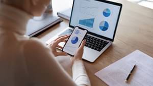 geschaeftsfrau-mit-smartphone-in-der-hand-sitzt-am-laptop-und-prueft-finanzuebersicht-mit-grafiken-auf-den-Bildschirmen