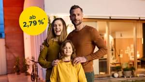 Familie steht glücklich mit dem Rücken zu ihrem renoviertem Haus und schaut in die Kamera: Modernisierungskredit ab 2,79% p.a.*