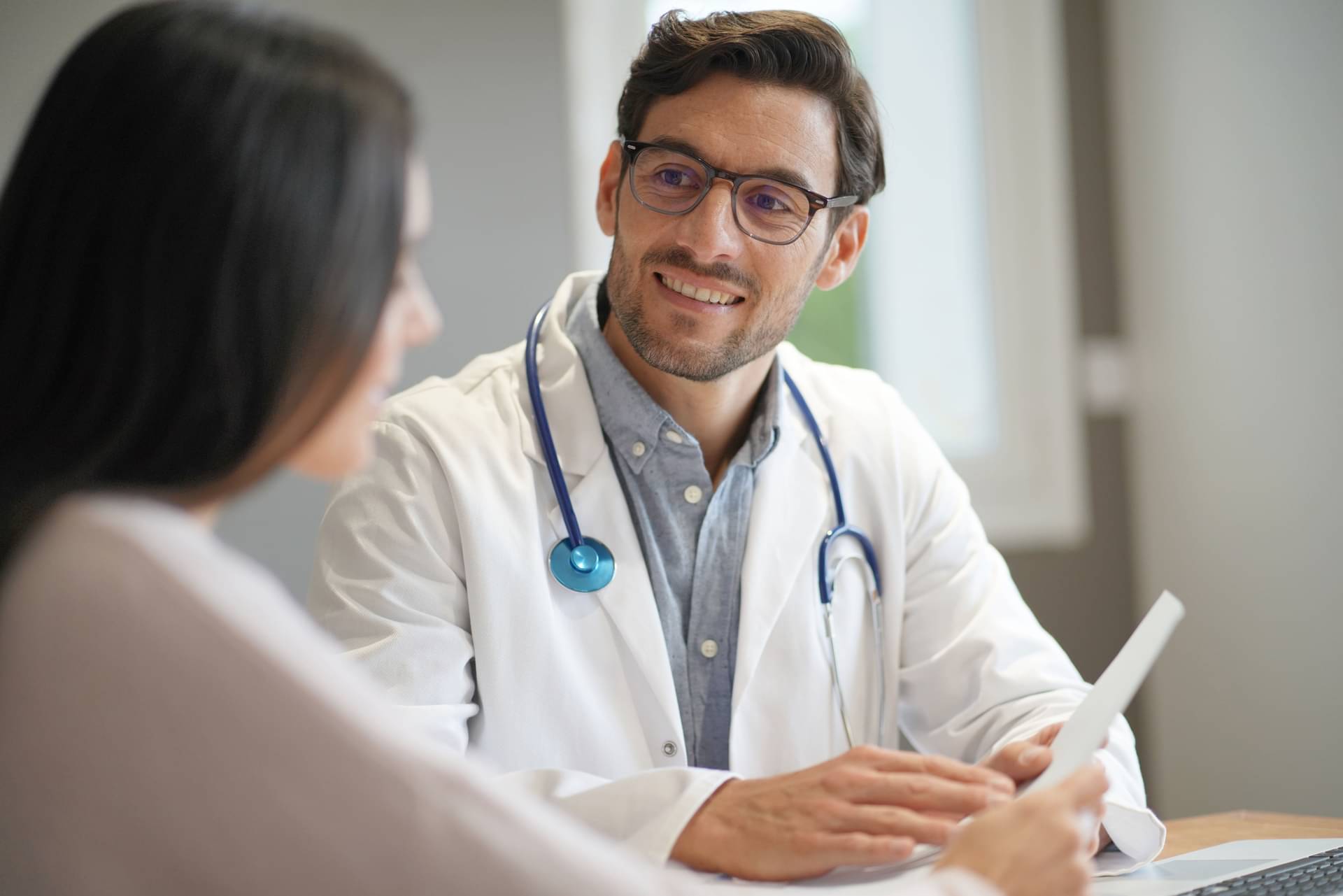 Ein lächelnder Arzt in Kittel und mit Stethoskop berät eine Patientin.
