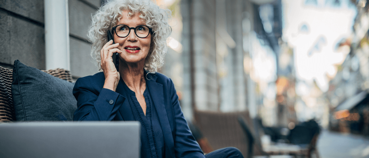 Mittelalte Frau mit grauen Haaren und Brille sitzt auf einem Sofa und träft einen blauen Blazer