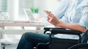 Frau sitzt an einem Schreibtisch im Rollstuhl und hat ein Tablet in der Hand
