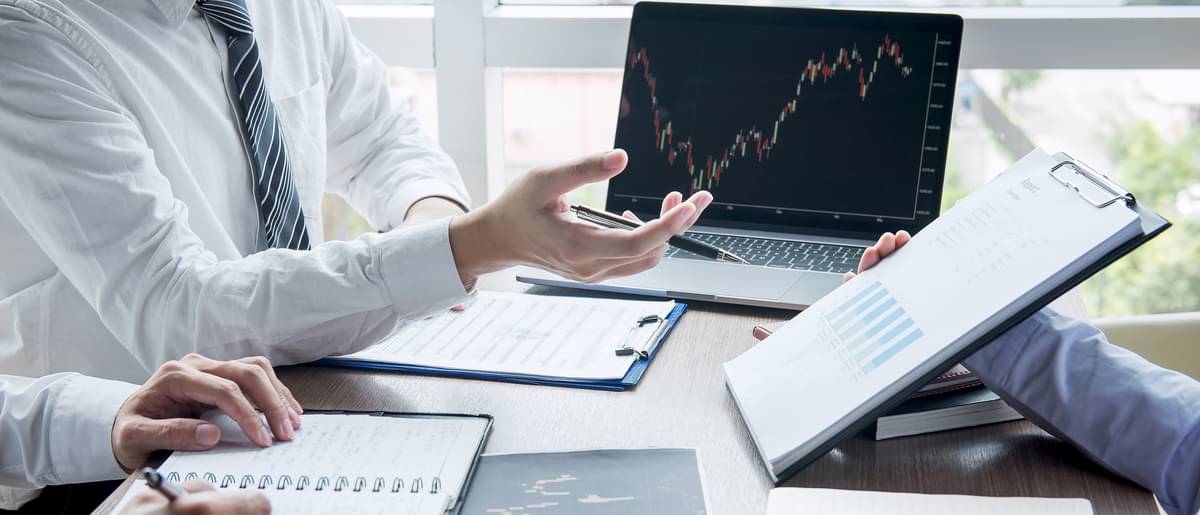 Besprechung zur Anlagestrategien, drei Personen sitzen am Schreibtisch vor Analogen und digitalen Unterlagen