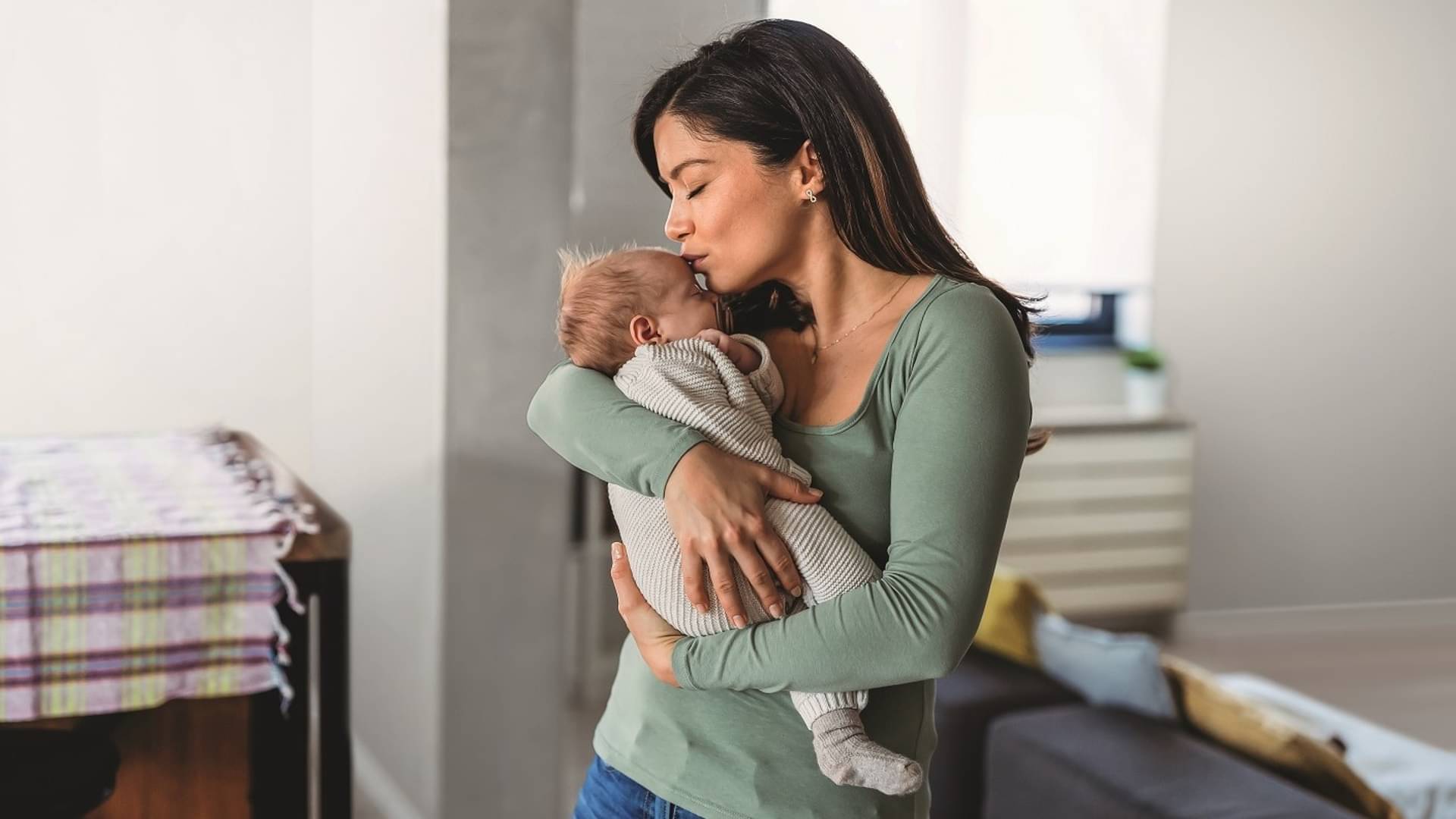 Eine junge Mutter hält ihr Baby im Arm und küsst dieses liebevoll auf die Stirn.
